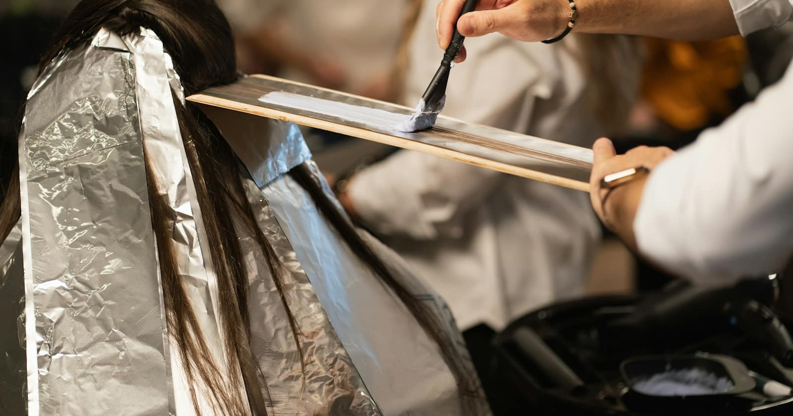 A stylist works on a client's colour in a salon while a phone lights up unanswered on the counter.
